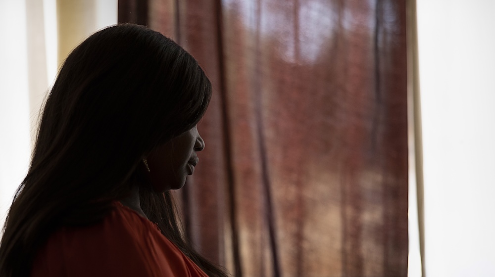 A woman stands before a curtained window with her face obscured. 