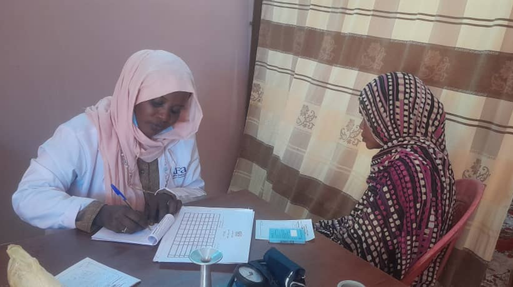 A woman sits across the table from a female health worker taking notes