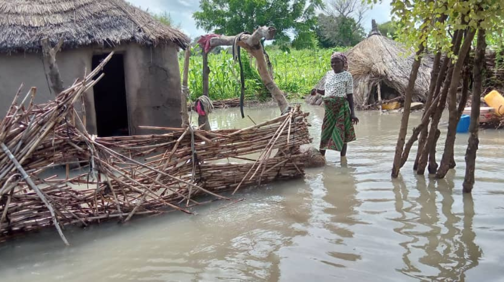 Una mujer rodeada de agua junto a una puerta de madera derrumbada y una casa con techo de paja