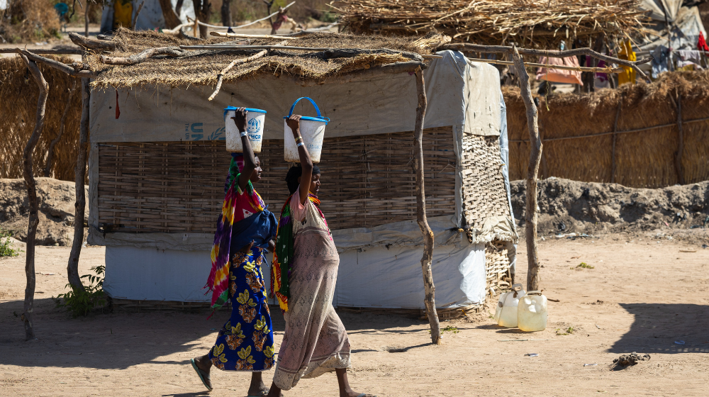 Two women walk past a makeshift shelter, holding UNHCR buckets on their heads