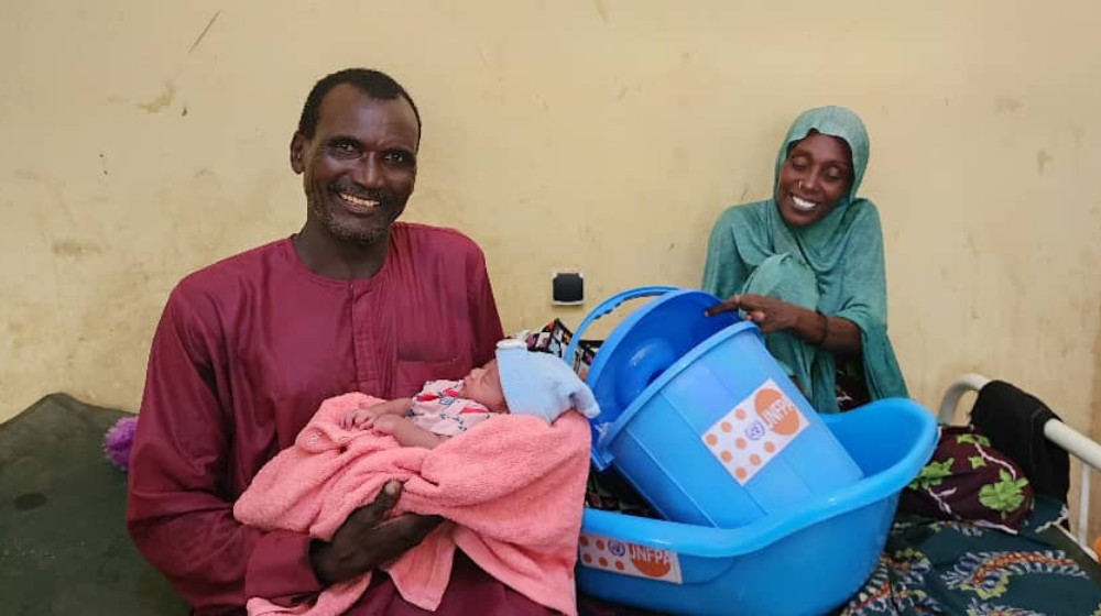 Un hombre vestido de rojo carga a un bebé recién nacido. Detrás, sentada en una cama, la madre del bebé, vestida de azul, sostiene una bañera de plástico azul con el logotipo del UNFPA.