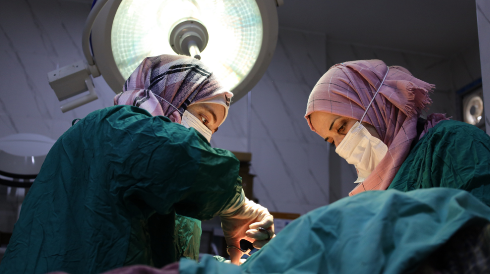 Two women medical workers wearing pink hijab and green operating gowns perform an operation under a large surgical light.