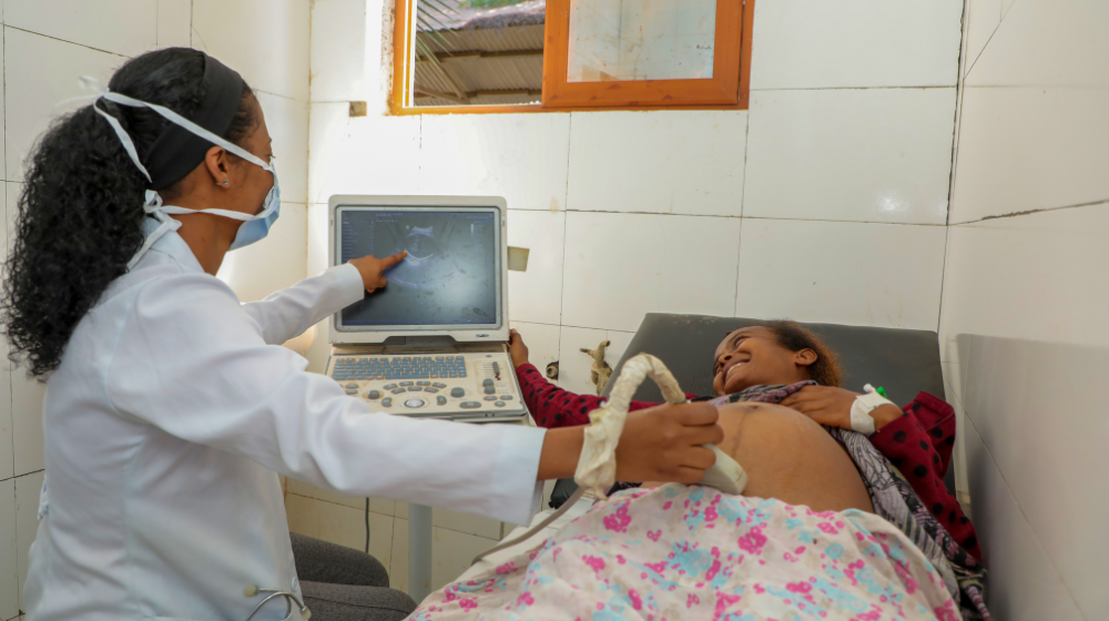 Una trabajadora sanitaria con bata blanca y mascarilla azul realiza un examen de ultrasonido a una sonriente mujer embarazada tumbada en una cama