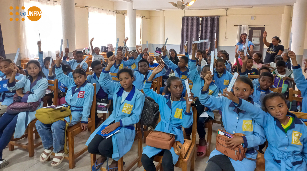 Un groupe de jeunes élèves en uniformes bleus lèvent la main, assis·e·s dans une salle de classe