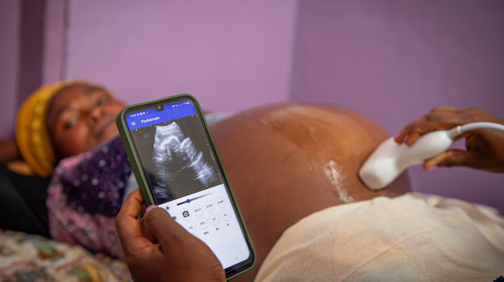  A woman in a yellow headwrap receives an ultrasound scan in a purple exam room. The ultrasound displays an image of a fetus on the midwife’s mobile phone