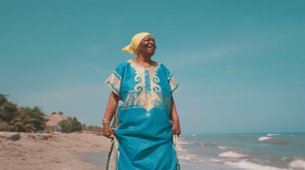 Une femme âgée d’ascendance africaine marche sur la plage, vêtue d’une robe bleue.