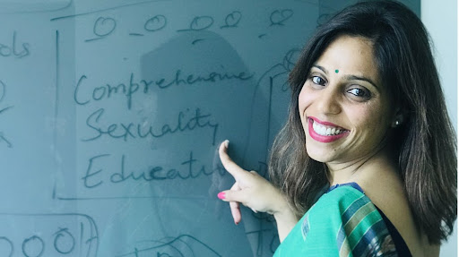 A woman smiles and points at a green board with her finger. 