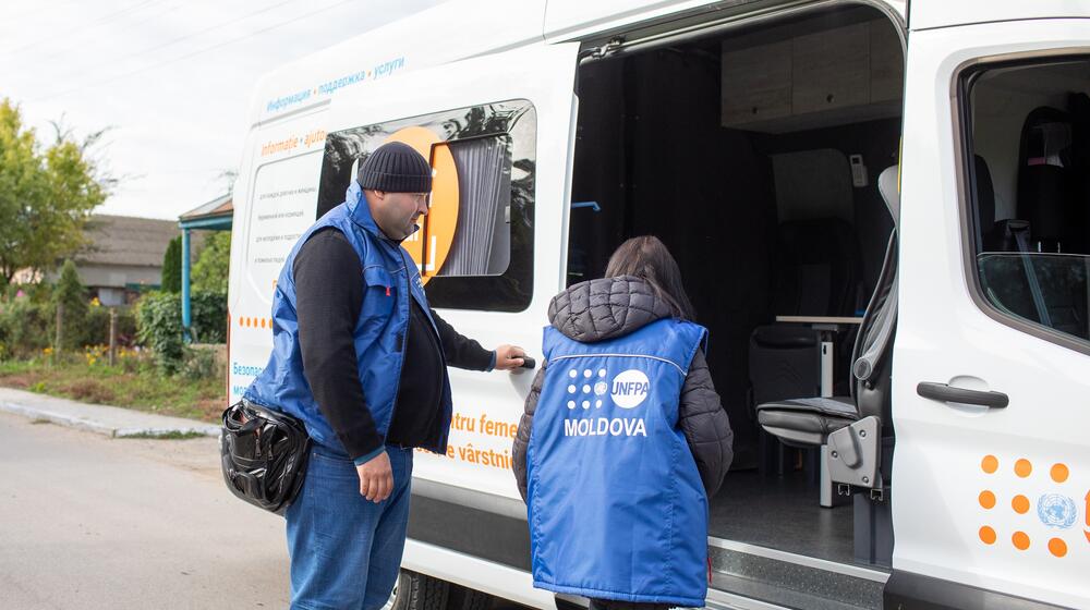 Two UNFPA workers stand next to a large mobile safe space vehicle which includes seating and a table.