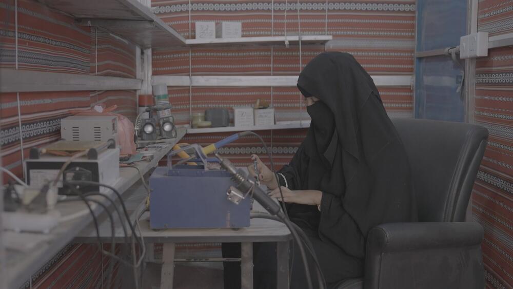 Une femme en niqab et abaya noirs est assise à un plan de travail, des outils à la main