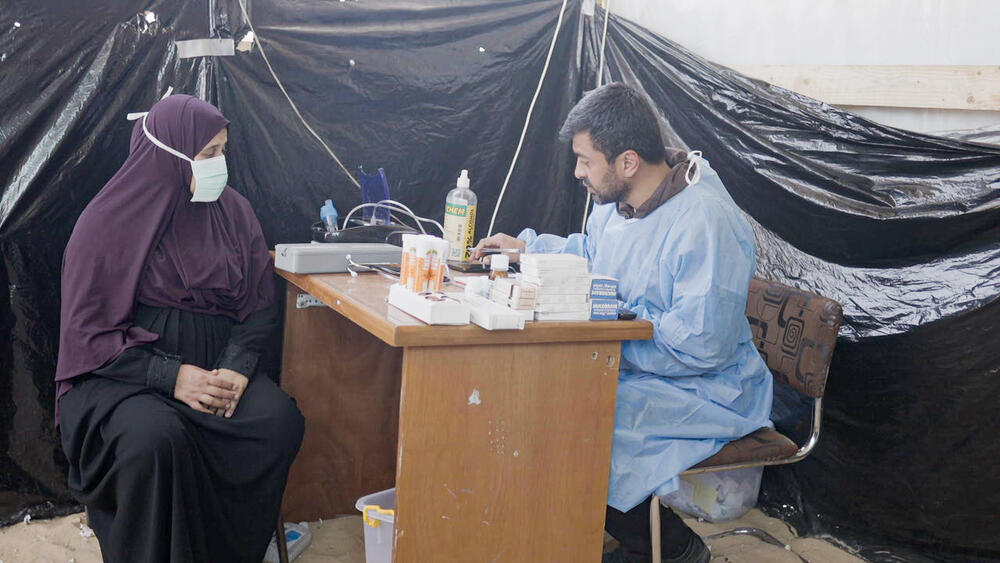 A man in blue scrubs sits at a desk in the sand, speaking with a pregnant woman