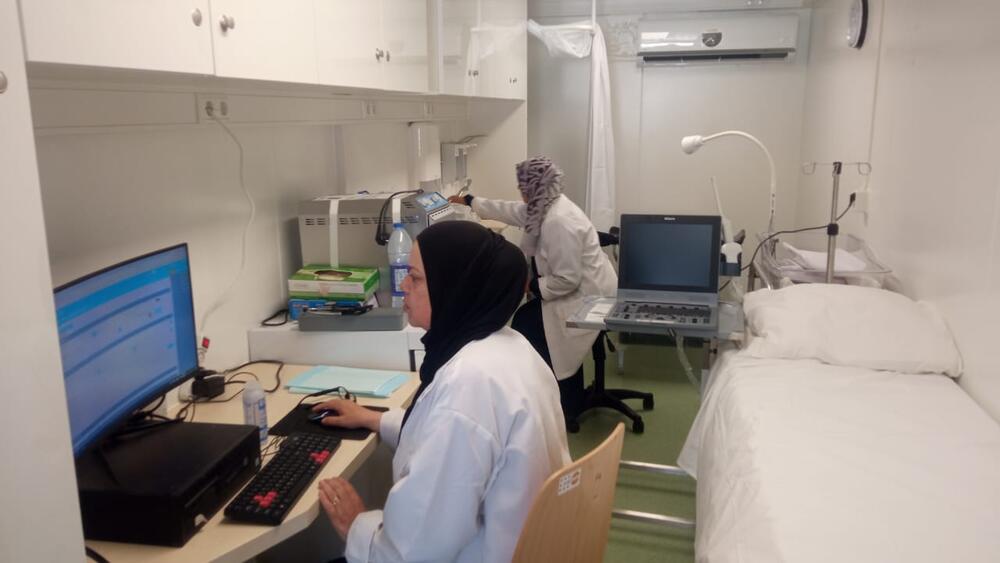 Two female health workers sit by an empty bed, one looking at a computer screen, the other taking a reading from a machine