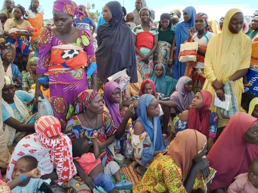 A group of women and babies huddle together outside, some seated, some standing.