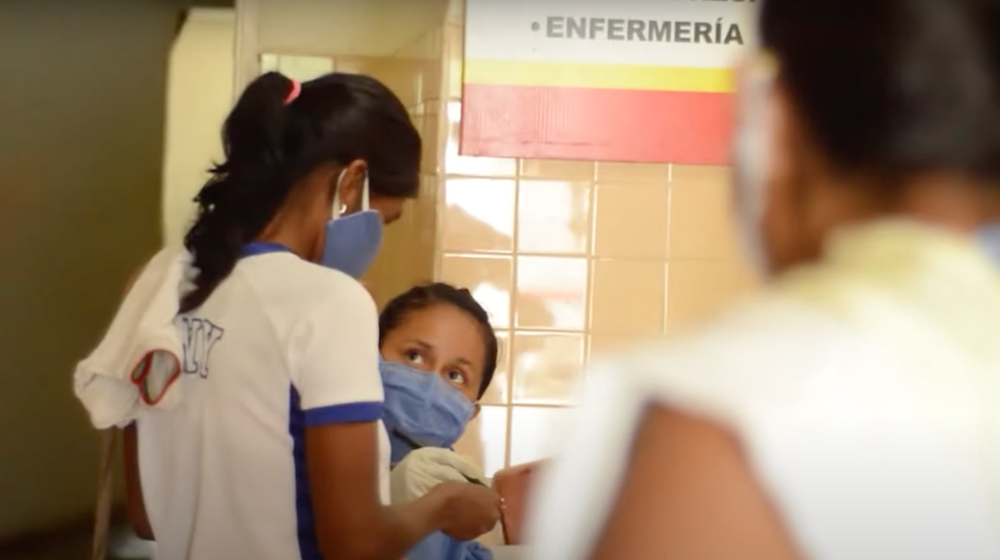 A female health worker talks to a female patient, both are wearing blue surgical masks