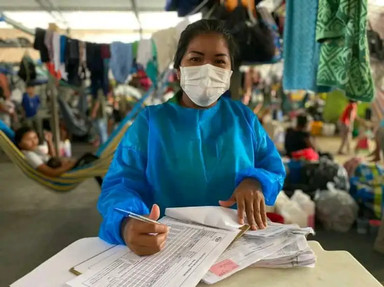 Una monitora de salud con bata y mascarilla trabaja en unos documentos.