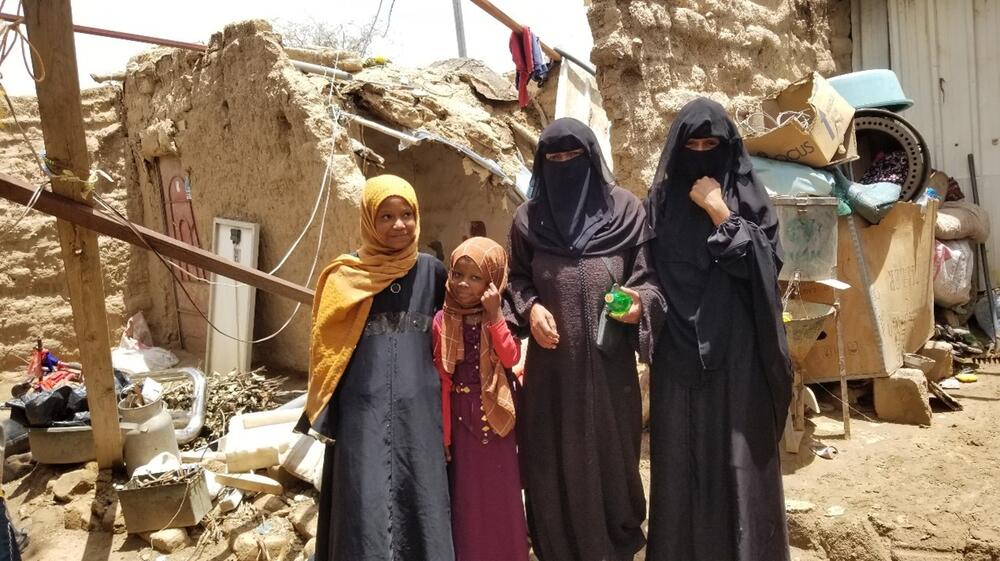 A family of women and girls stands outside of a damaged shelter.