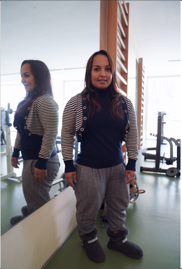 A young woman stands in a fitness centre, looking straight ahead and smiling, reflecting in a large mirror to her right. 