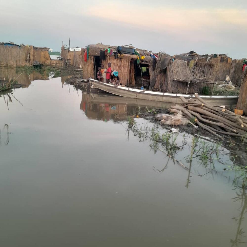  Une famille se tient dans l’entrée d’une maison à toit de chaume. On voir leur reflet dans l’eau qui entoure leur maison.