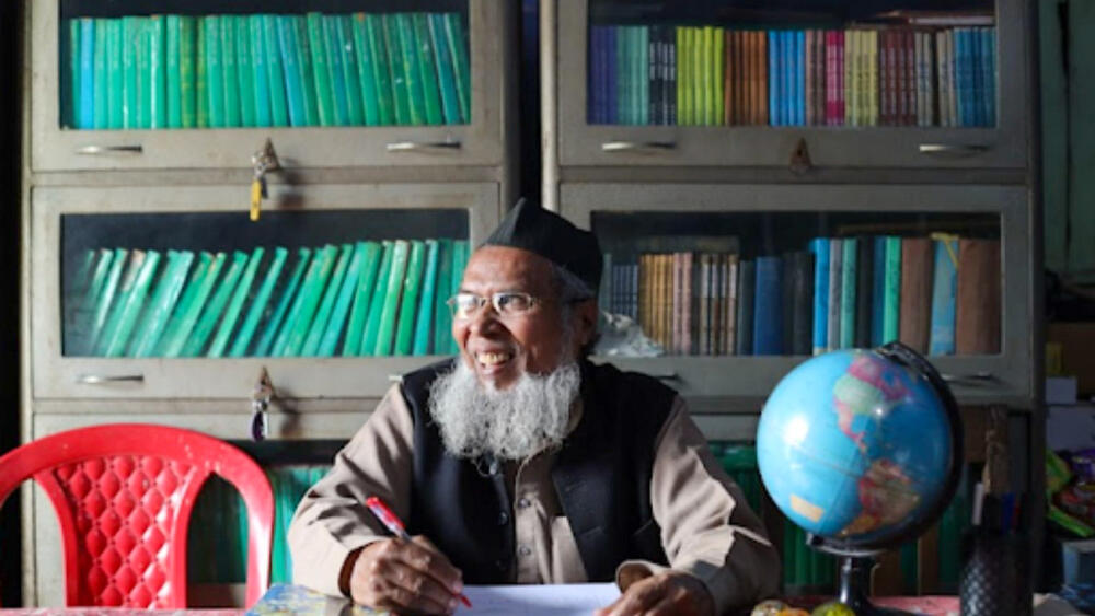 A man holding a pen sits at a desk in front of a bookshelf.