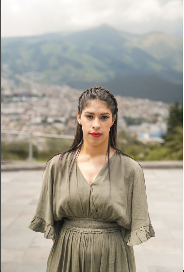 A young woman wearing red lipstick stands in a plaza, looking straight ahead and half-frowning, half smiling.