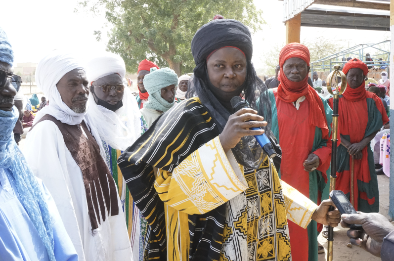 A village chief, surrounded by other men, speaks into a microphone.