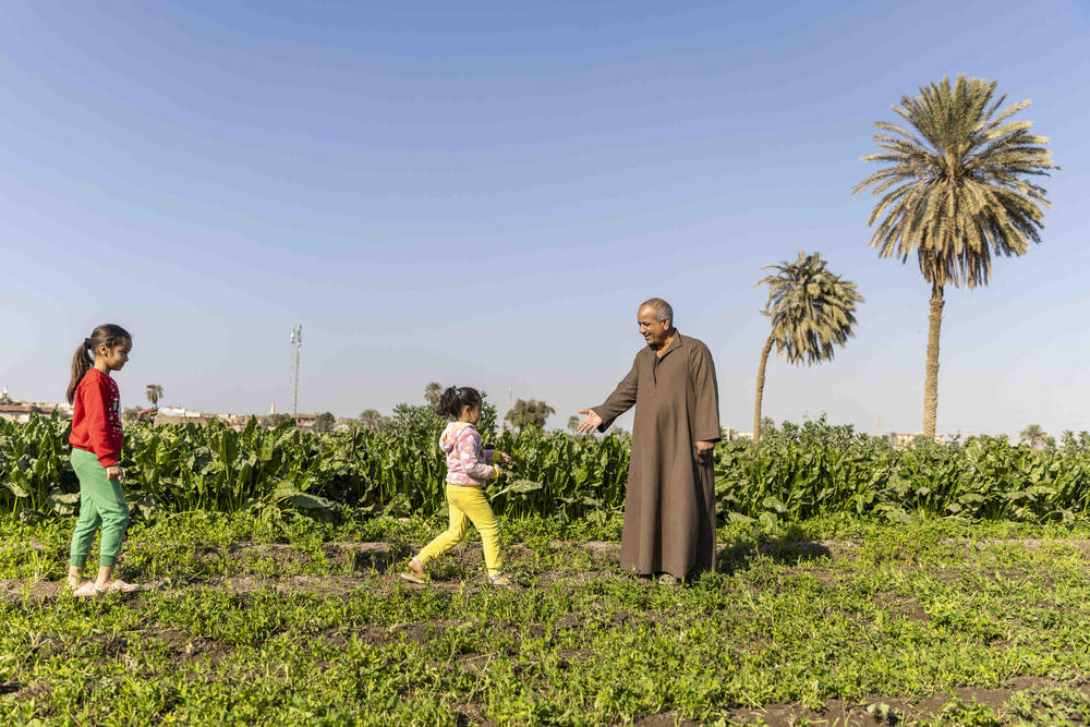 رجل يرتدي سترة بنية اللون يمد يده لفتاتين صغيرتين تسيران نحوه. ويمكن رؤية شجرتي نخيل ونباتات خضراء خلفهما.