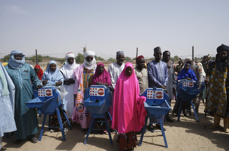 Des adolescentes se tiennent en compagnie d’anciens du village près de broyeurs à grain portant le logo de l’UNFPA