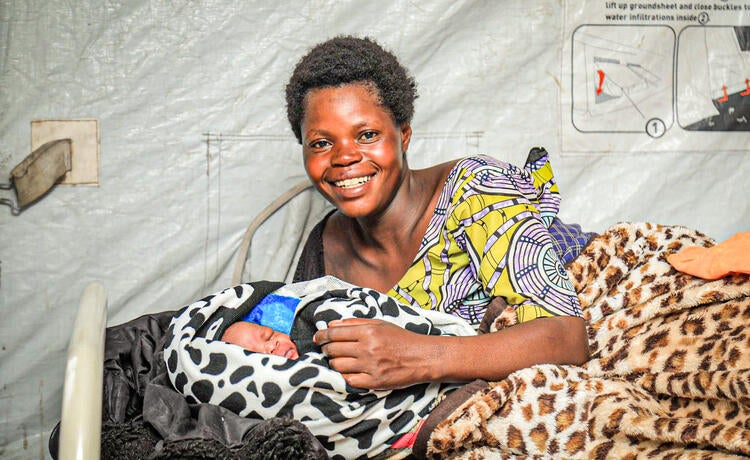 A woman lying on a bed inside a tent cradles her newborn baby while smiling into the camera