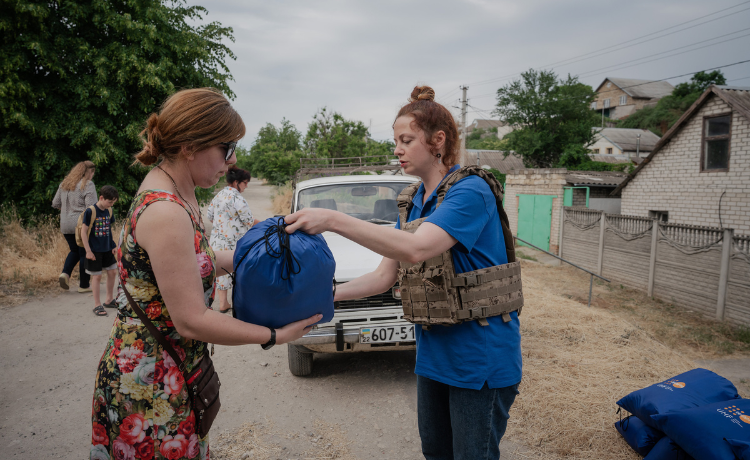 : A woman in a combat vest hands a blue bag of UNFPA supplies to a woman in a flowery dress]