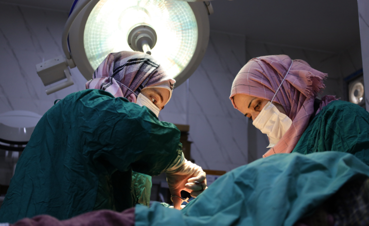 Two women medical workers wearing pink hijab and green operating gowns perform an operation under a large surgical light.