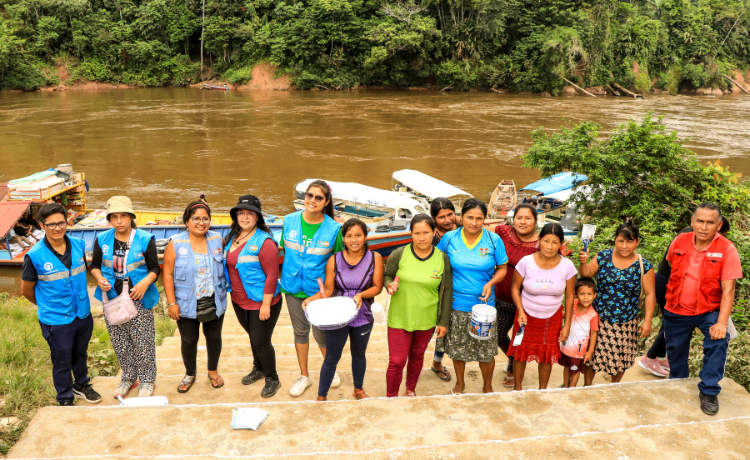 A group of fourteen people looking at the camera. Some have blue UNFPA-branded vests, and others hold buckets with white paint. They are all standing at the harbour steps surrounded by the jungle.