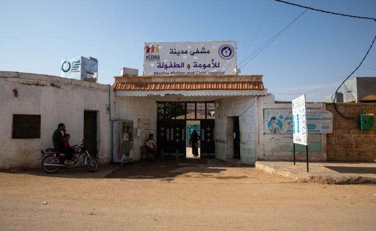 El exterior del Hospital de Maternidad y Neonatología Medina, con un hombre y un niño esperando en una motocicleta y una mujer avanzando por la entrada