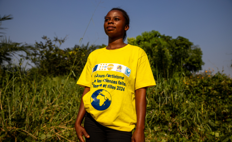 A woman in a yellow t-shirt stands in front of green vegetation smiling