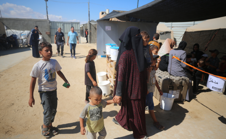 A pregnant woman holds the hand of a young child, followed by an older child as she walks through the sand beside tens. Some other people are visible i the background