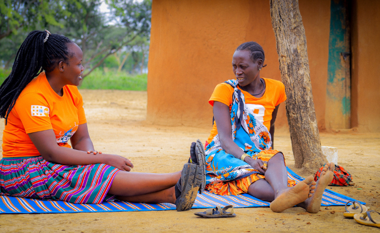 Una mujer con camiseta naranja con el logotipo del UNFPA habla con la Sra. Telo, que también lleva una camiseta naranja. Están sentadas en una manta a rayas en un patio.