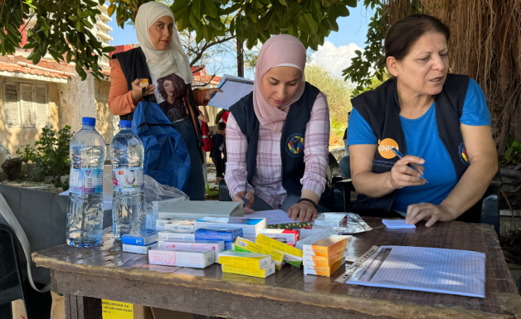 Women arrange medicines and supplies on a table under a tree. They are wearing black vests with the logos of UNFPA and Mosaic.