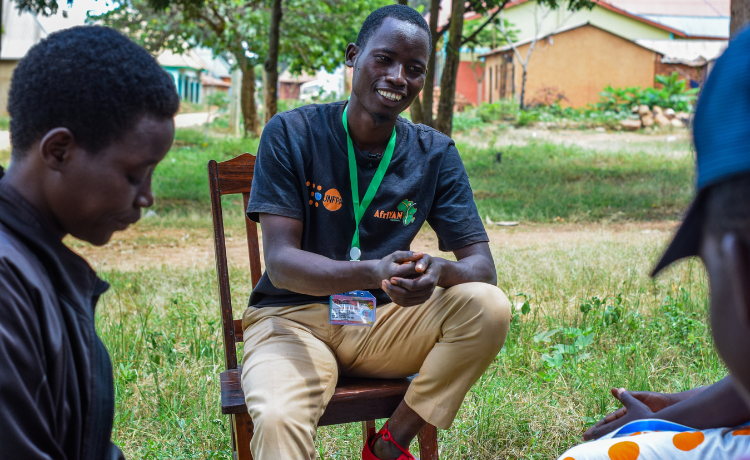  A man in a black t-shirt with an orange UNFPA logo sits on a chair in the shade of a courtyard. He is speaking to other young pرجل يرتدي قميصًا أسود عليه الشعار البرتقالي لصندوق الأمم المتحدة للسكان يجلس على كرسي في الظل في فناء. إنه يتحدث إلى شباب آخرين.eople.