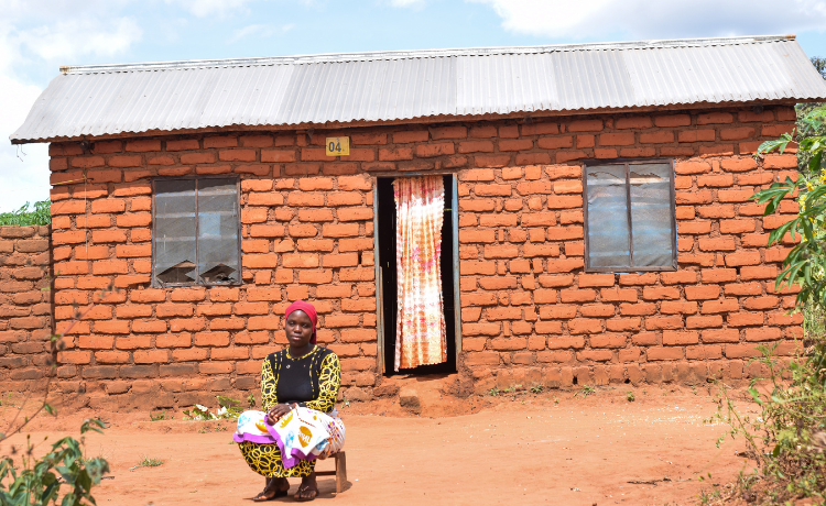 Une femme portant un foulard rouge sur la tête est assise devant une maison en briques, au toit en métal ondulé