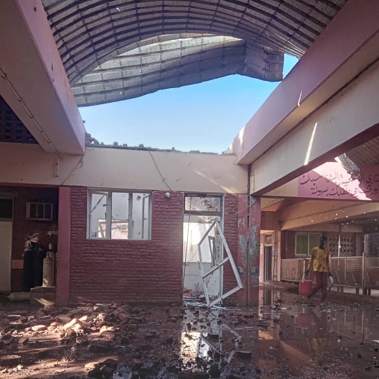 A person in a yellow t-shirt carries a red bucket through rubble and puddles. The ceiling is missing.