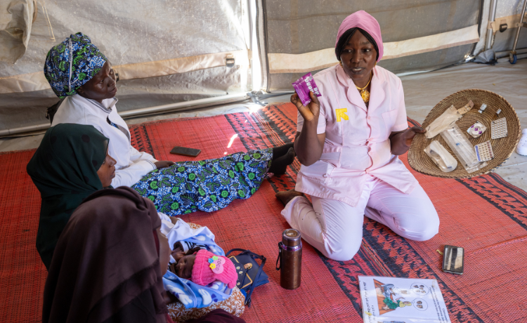 Une sage-femme en blouse rose est assise par terre dans une tente ; elle présente à trois autres femmes assises un plateau tissé sur lequel sont disposés des contraceptifs