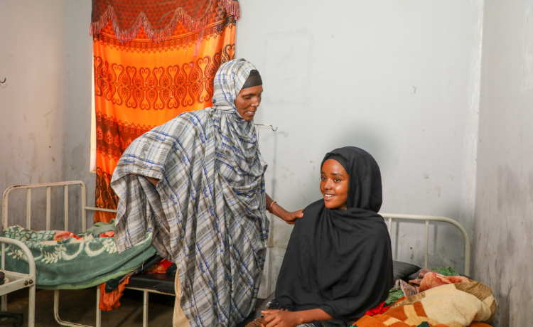 A young mother sits on a hospital bed beside her newborn, an older woman stands beside her, smiling with her hand on the new mother;s shoulder.