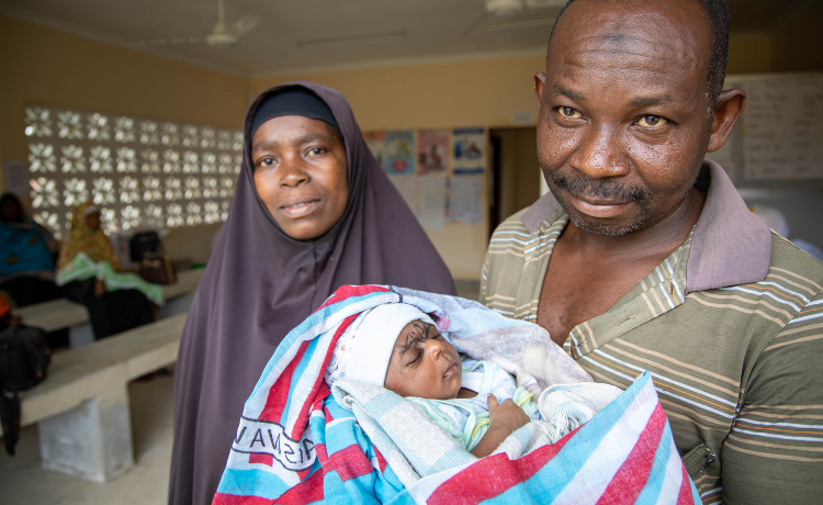 A man in a striped polo shirt holds his infant child, who is wrapped in a blanket. The child’s mother, wearing a black hijab, stands to the side smiling.