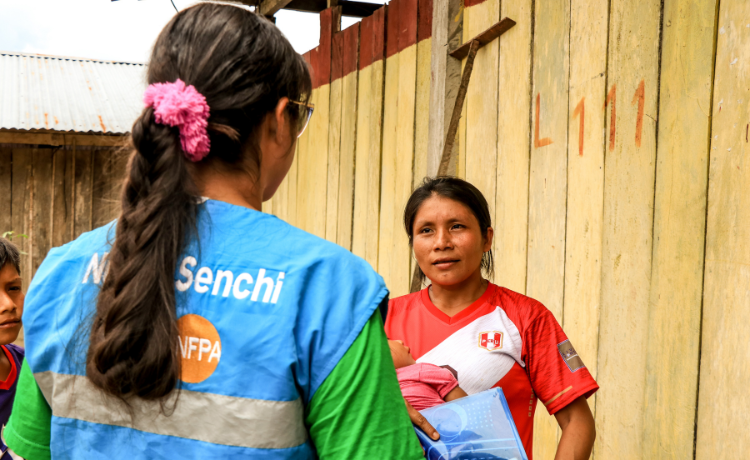 Two women outside a wooden home. One of them has a UNFPA-branded blue vest, the other holds a small child