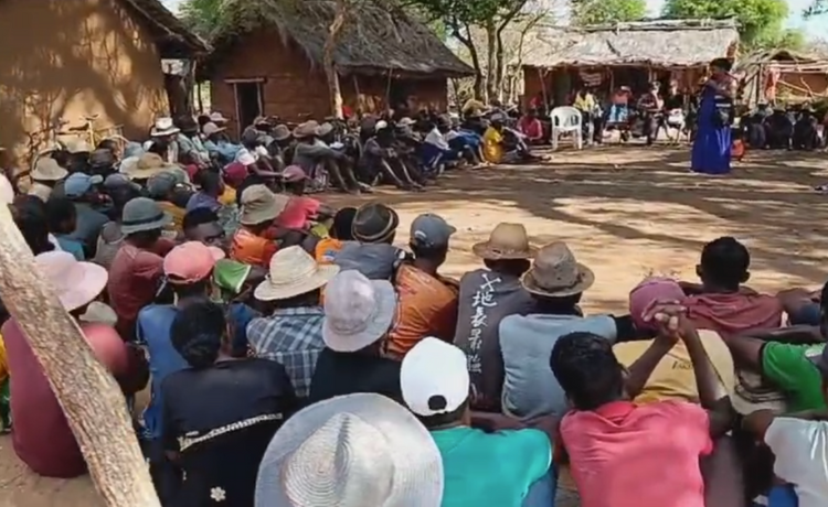 A group of people seated around a clearing in a village listen to a woman speaking in the middle