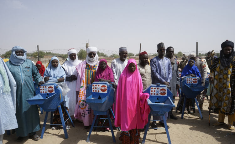 Teenage girls and village elders stand beside seed grinding mills with the UNFPA logo