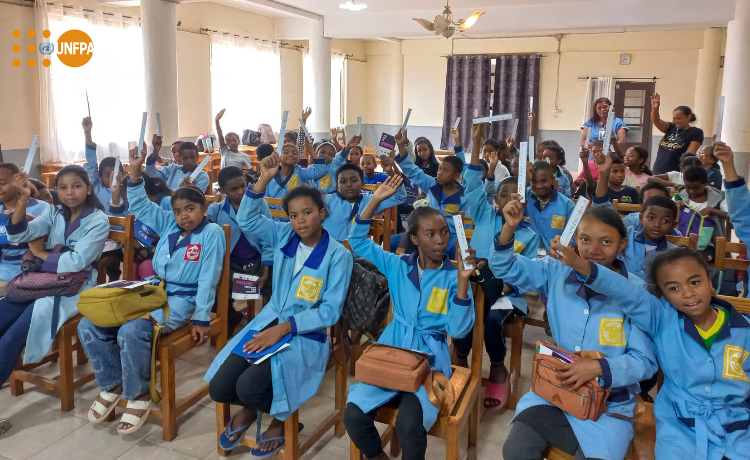 Un grupo de jóvenes estudiantes con uniforme azul levantan la mano sentados en un aula