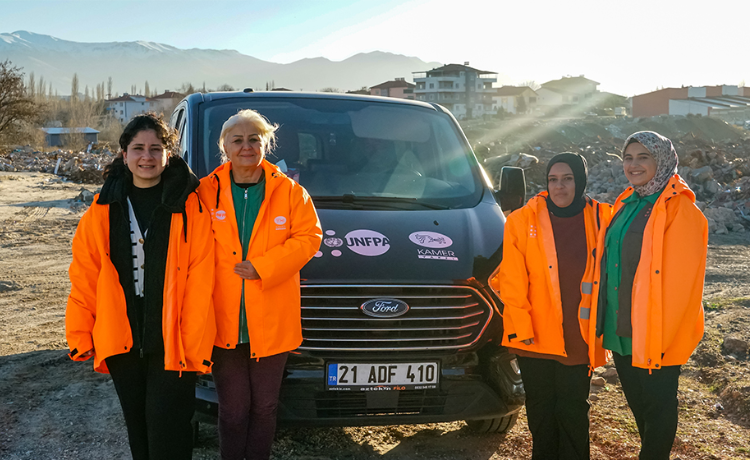  Cuatro mujeres con brillantes chaquetas color naranja de pie frente a un automóvil en medio de escombros causados por el terremoto de 2023 en Türkiye.