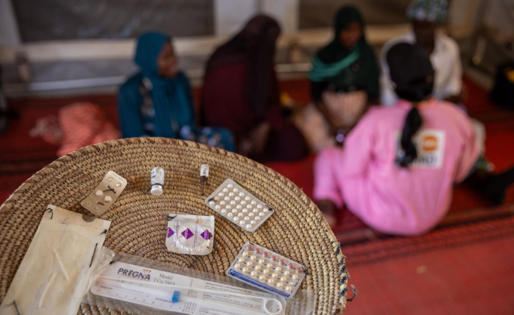 À l’arrière-plan, une agente de santé en uniforme rose est assise sur le sol et s’adresse à un groupe de femmes ; au premier plan, un panier tressé contient divers contraceptifs modernes