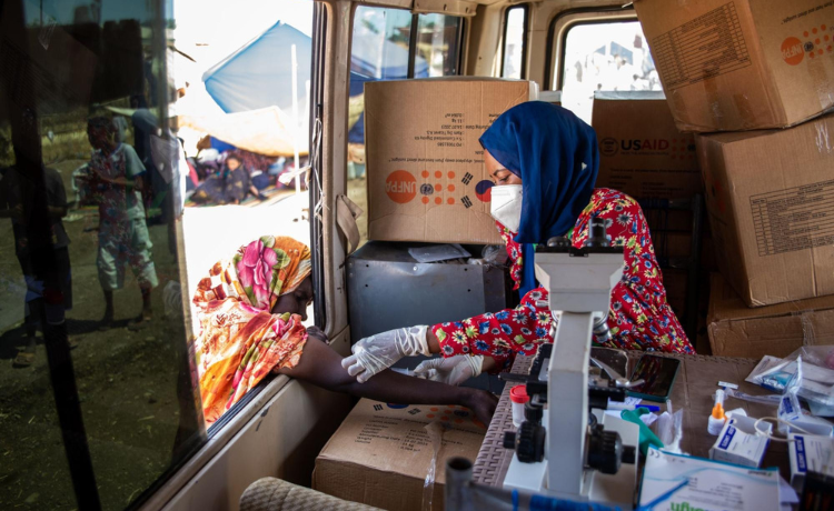 Una mujer enmascarada y con velo sentada en una camioneta tras un microscopio y frente a cajas etiquetadas por el UNFPA administra medicamentos al brazo de una mujer mientras se inclina por la ventana abierta.