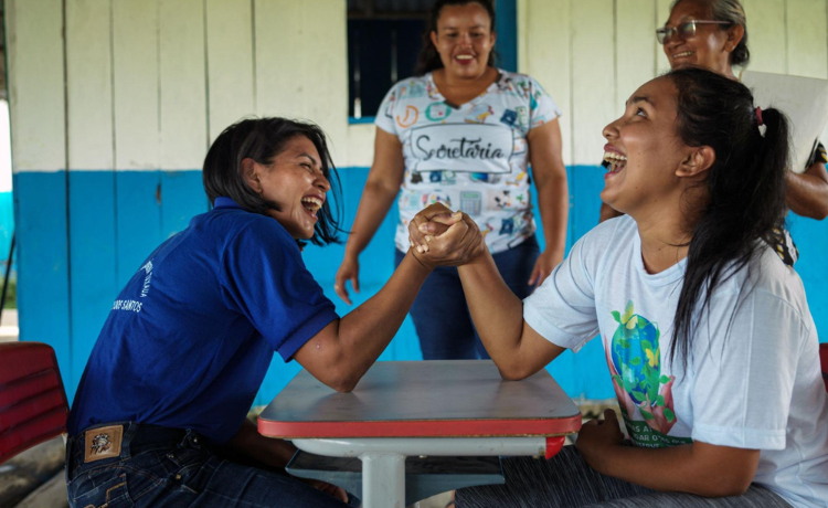 Dos mujeres en una mesa pulsean, riendo, mientras otras dos mujeres miran sonriendo al fondo