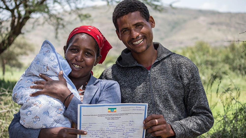 A father and mother hold up a printed paper, a newborn is in the mother's left hand.